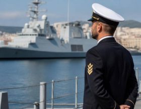 Militare della Marina in uniforme blu e berretto bianco sul ponte di una nave ormeggiata, con strutture navali sullo sfondo.