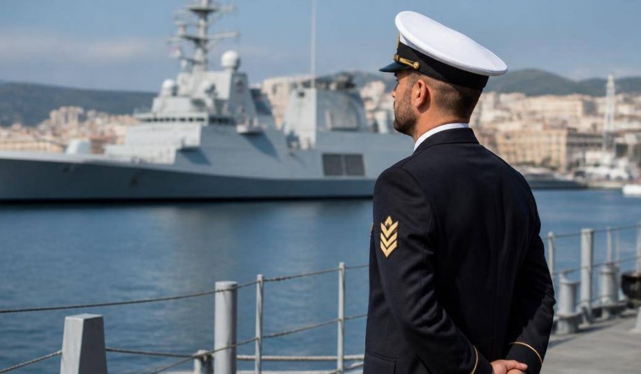 Militare della Marina in uniforme blu e berretto bianco sul ponte di una nave ormeggiata, con strutture navali sullo sfondo.
